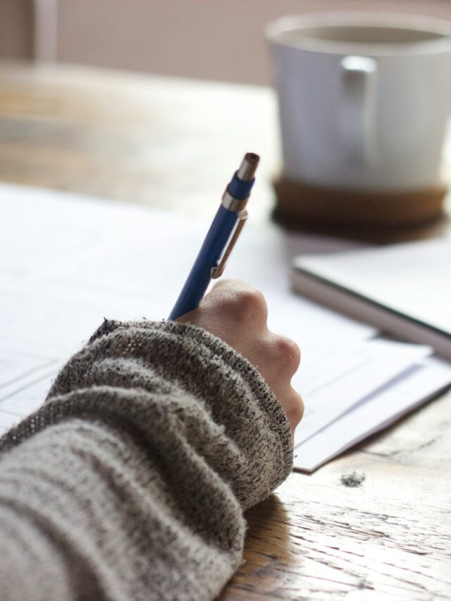 person writing on brown wooden table near white ceramic mug