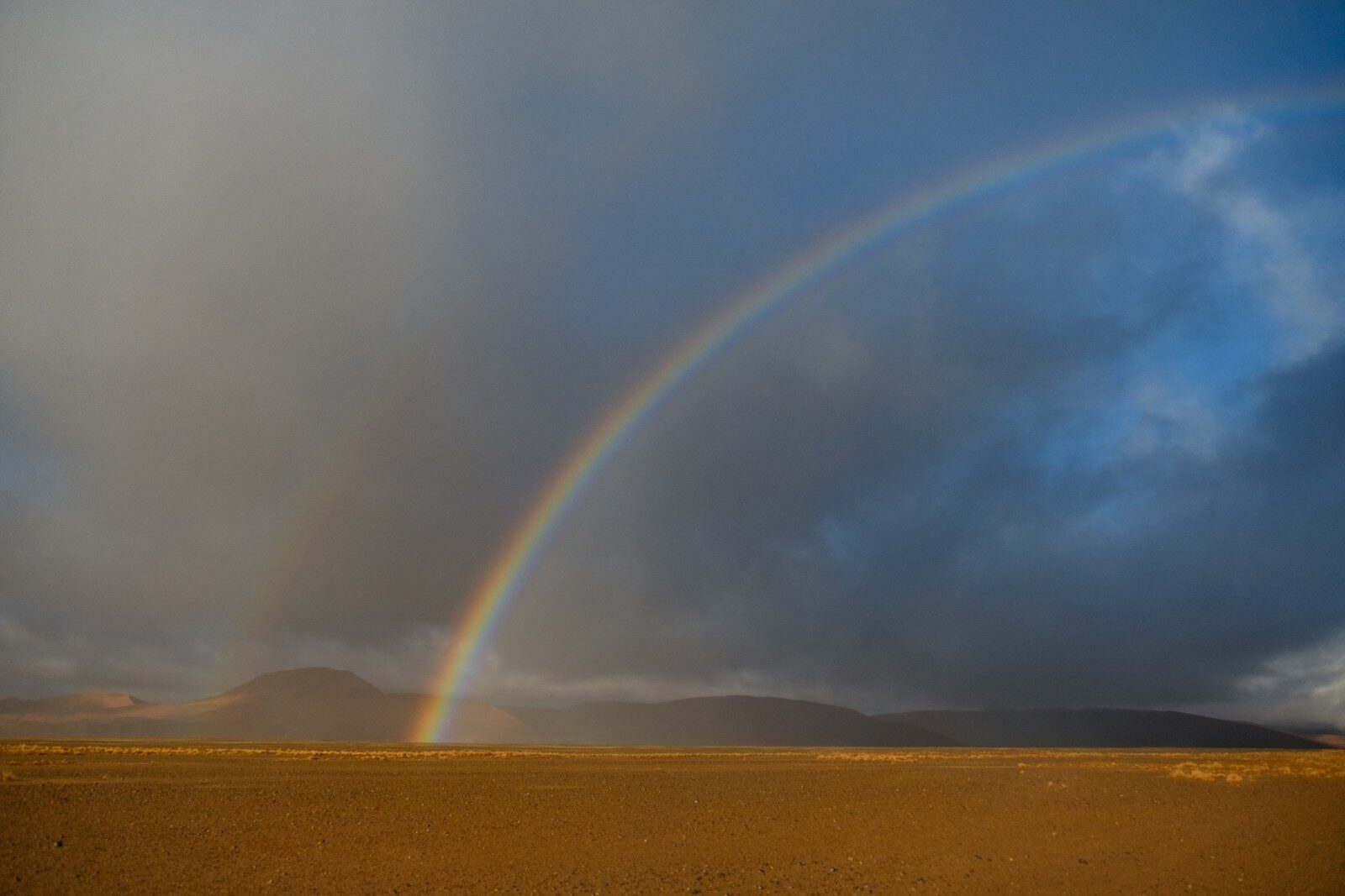 A vibrant rainbow arching over a vast desert landscape under a dark cloudy sky