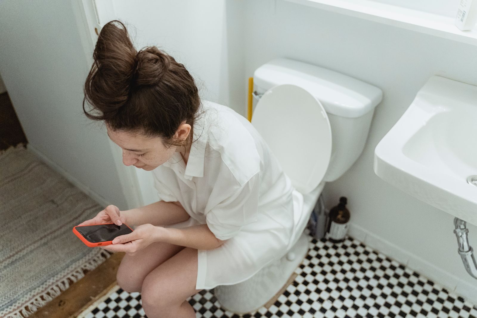 A woman sitting in a bathroom, focused on her smartphone, from a high angle view.