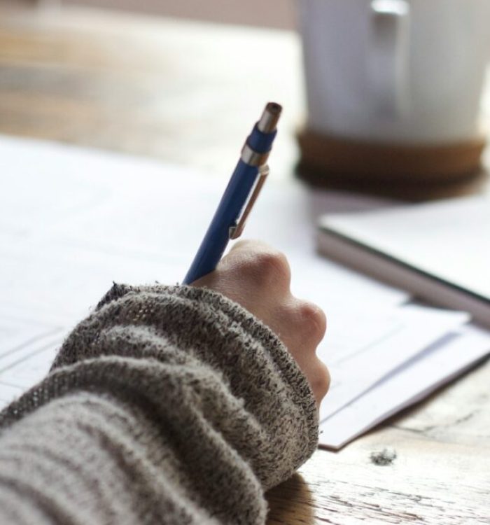 person writing on brown wooden table near white ceramic mug